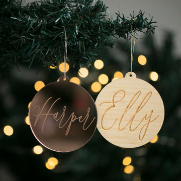Two Script Christmas Baubles with the names “Harper” and “Elly” engraved in cursive hang from a tree branch, with softly blurred yellow holiday lights glowing in the background.