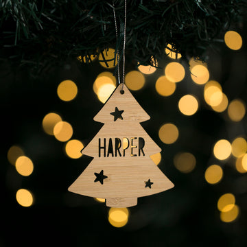 A personalised Evergreen Christmas Tree ornament in bamboo hangs from a string, featuring the name Harper and star cutouts, with blurred warm yellow lights glowing softly in the background.