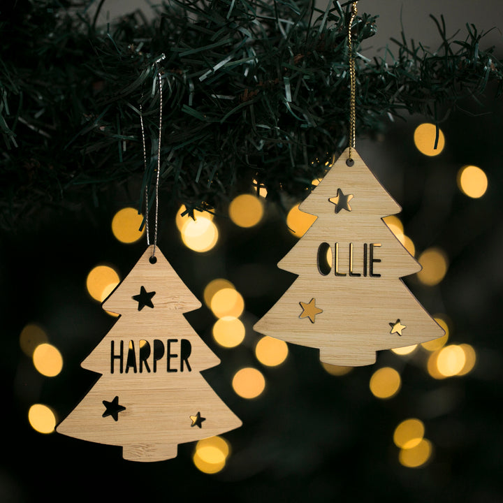 Two Evergreen Christmas Tree ornaments hang from a branch. Each bamboo bauble, engraved with a name—one HARPER, the other OLLIE—features star and circle cut-outs, with blurred lights shining in the background.