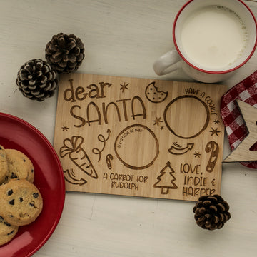 A festive scene with a Personalised Easter Bunny Treat Board for cookies, milk, and a carrot, surrounded by pinecones, chocolate chip cookies on a plate, and a mug of milk atop a white table with a red and white checked napkin.