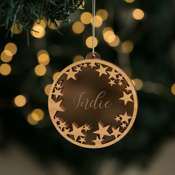The Oh Starry Night Christmas Bauble, featuring star cutouts and ‘Indie’ engraved at the center, hangs from a tree branch with softly glowing holiday lights blurred in the background.