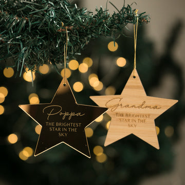 Two Brightest Star Memory Christmas Baubles hang from a green Christmas tree. One reads Poppa THE BRIGHTEST STAR IN THE SKY, the other Grandma THE BRIGHTEST STAR IN THE SKY. Soft, blurred lights shine in the background.