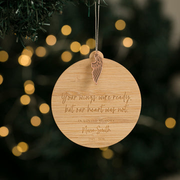 A round bamboo Christmas bauble with an engraved quote and wing design, hanging from a tree with holiday lights in the background.