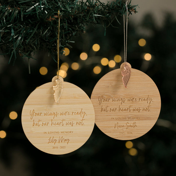 Two Memory Wings Christmas Baubles with angel wing charms hang on a tree. Each wooden ornament is personalised with a name, dates, and message. Softly glowing lights create a warm background.