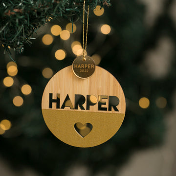 A Keepsake Disc for Baubles (bauble not included) hangs on a tree, with HARPER cut out above a heart and a gold disc engraved HARPER 2022. Soft holiday lights glow behind this charming personalised decoration.
