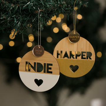 Two Keepsake Discs for Baubles (bauble not included) hang on a tree, one reading “INDIE 2022” with a heart cutout, the other “HARPER 2022,” glowing softly amid holiday lights—perfect personalised Christmas decorations.