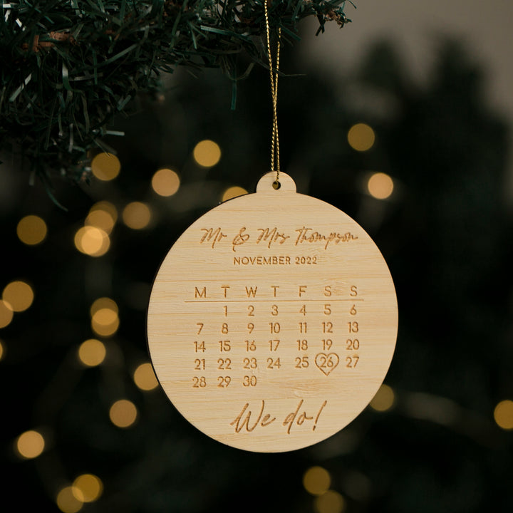 The Perfect Date Christmas Bauble, a wooden calendar-shaped decoration with "Mr & Mrs Thompson," "November 2022," a heart on the 26th, and "We do!" engraved, makes a thoughtful personalised engagement gift for the tree. Blurred holiday lights glow behind.