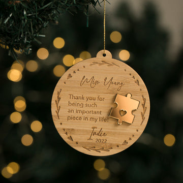 A personalized Christmas bauble with a puzzle piece shape cut out, hanging from a golden twine. It has an inscription thanking a teacher and is placed on a Christmas tree with lights.
