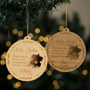 Two round wooden Puzzle Piece Teacher Christmas Baubles hang on a tree, each engraved with names, a puzzle piece design, and the message: “Thank you for being such an important piece in my life!” with personalised names and dates.