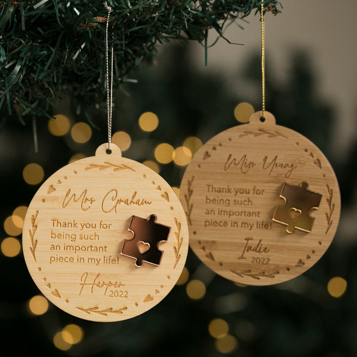 Two round wooden Puzzle Piece Teacher Christmas Baubles hang on a tree, each engraved with names, a puzzle piece design, and the message: “Thank you for being such an important piece in my life!” with personalised names and dates.