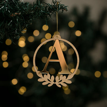 The Laurel Letter Christmas Bauble, a gold initial ornament featuring the letter A, hangs from a tree branch against softly blurred yellow lights.