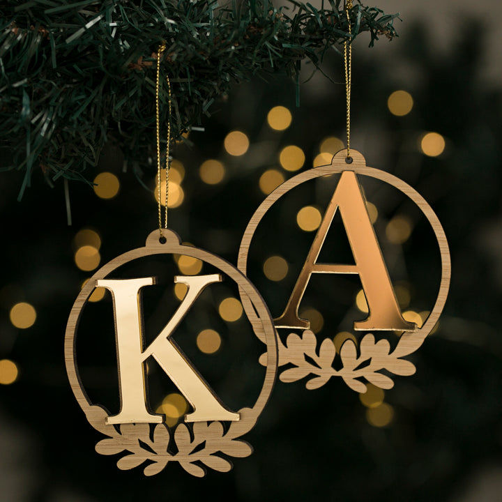 Two Laurel Letter Christmas Baubles, circular wooden ornaments with cutout K and A, hang from a tree branch with gold string and blurred festive lights—a charming personalised touch for your holiday décor.