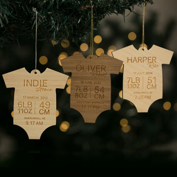 Three Baby Bodysuit Christmas Bauble ornaments engraved with birth details hang on a tree, fairy lights glowing behind. These personalised keepsakes are a charming gift to celebrate baby’s first Christmas.