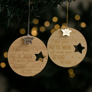 Two Infinity Christmas Baubles with star cutouts hang on a Christmas tree. Each personalised ornament is engraved with “I love you past the moon & miss you beyond the stars” and features names like “Winston” or “Dad” on each star.