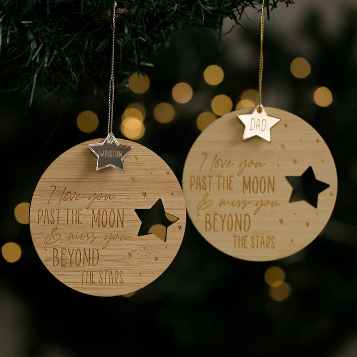Two Infinity Christmas Baubles with star cutouts hang on a Christmas tree. Each personalised ornament is engraved with “I love you past the moon & miss you beyond the stars” and features names like “Winston” or “Dad” on each star.