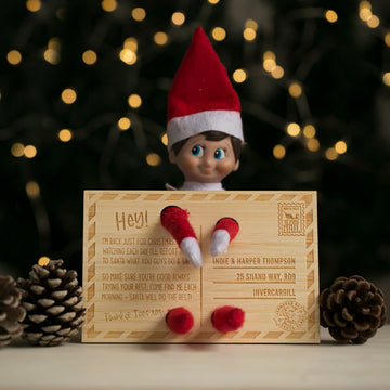 The Elf on the Shelf Arrival Postcard features a toy elf with a red hat holding a personalised card with red felt arms and legs, surrounded by pinecones and festive lights—a charming accessory from our Christmas range.