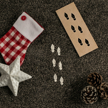 An Elf Footprints Stencil, Christmas stocking, white ceramic star, and pinecones rest on the carpet. White footprints created with the Elf Footprints Stencil add a whimsical touch to this festive tradition.