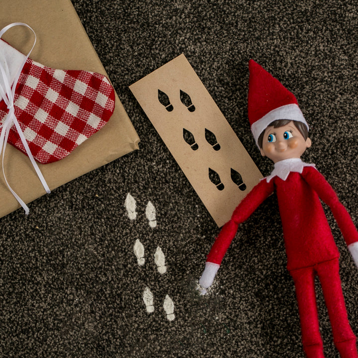 A red Elf on the Shelf doll lies on dark carpet next to an Elf Footprints Stencil, white painted footprints, a wrapped gift, and a red-checkered Christmas stocking—a fun scene from this cherished holiday tradition.