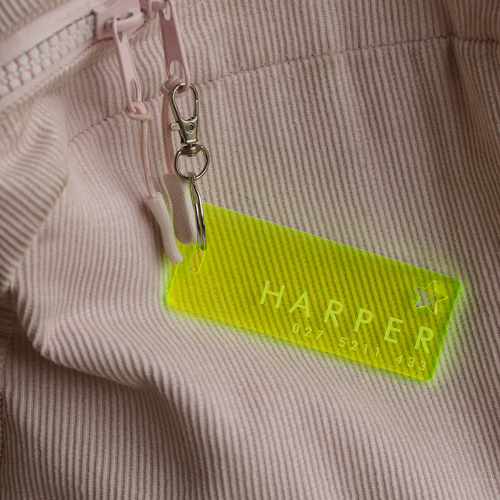 A neon yellow “HARPER” bag tag with a phone number, The Details Key Ring, is attached to the zipper of a light pink ribbed fabric bag.
