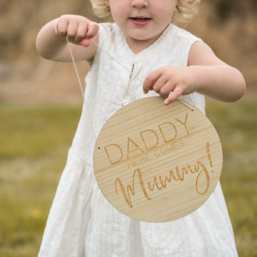 A girl holding a round light bamboo sign saying Daddy here comes Mummy!