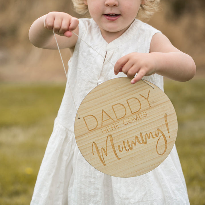 A girl holding a round light bamboo sign saying Daddy here comes Mummy!