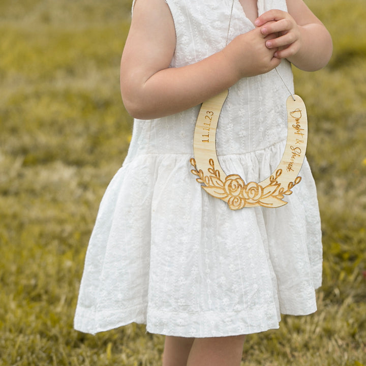 A young child in a white dress stands on grass, wearing the NZ-made Personalised Floral Wedding Horseshoe engraved with a date and floral design. Hanging from their neck, this unique wedding gift keeps the child&