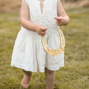 A young child in a white dress stands barefoot on grass, holding a Personalised Floral Wedding Horseshoe—a unique NZ-made gift decorated with flowers and custom writing. The child&