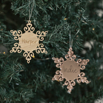 Two Personalised Christmas Snowflakes with intricate designs hang on a tree, each engraved with names like Harper and Indie, adding a unique touch to your festive décor.