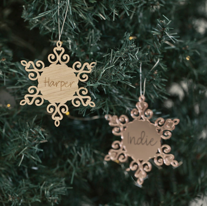 Two Personalised Christmas Snowflakes with intricate designs hang on a tree, each engraved with names like Harper and Indie, adding a unique touch to your festive décor.