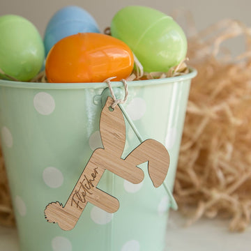 Mint green Easter bucket with a wooden F laser cut letter with Fletcher engraved into it. The letter has bunny ears and bunny tail and the bucket is full of bright colours plastic fillable eggs. Wood wool is in the background.