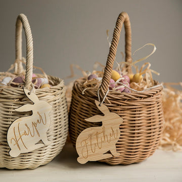 Two eco-friendly bamboo Easter baskets with straw and pastel eggs sit side by side. Each features a Personalised Vintage Easter Bunny Tag, handwritten with the names Harper and Fletcher, tied to its handle.
