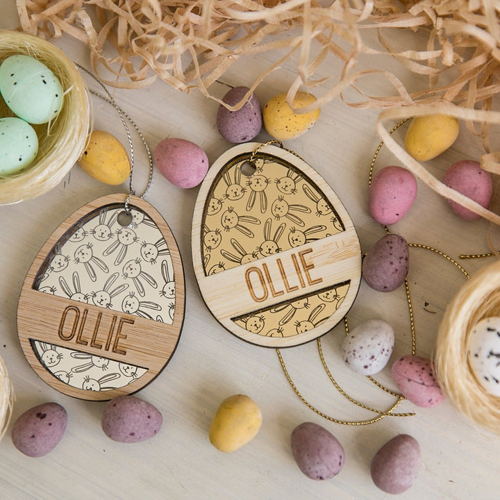 Two Personalised Golden Egg Easter Tags labeled OLLIE rest on a table with candy eggs, straw, and twine. These custom tags feature engraved bunny and chick designs—perfect as unique Easter Bunny gift tags.