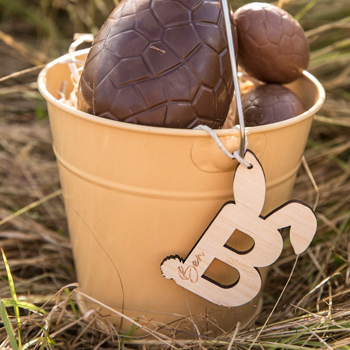 A beige bucket of chocolate eggs sits on dry grass. Attached to its handle is a Personalised Easter Letters eco-friendly bamboo tag—a wooden bunny engraved with the letter B and the name Ben.