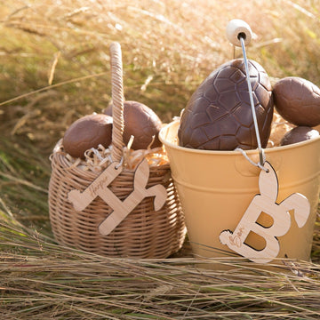 A wicker easter basket and a tin easter pale with engraved H and B bamboo letters, both with a bunny tail and bunny eggs. Chocolate eggs fill the baskets.