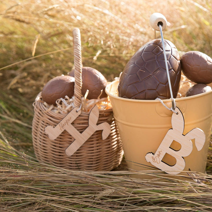 A wicker easter basket and a tin easter pale with engraved H and B bamboo letters, both with a bunny tail and bunny eggs. Chocolate eggs fill the baskets.