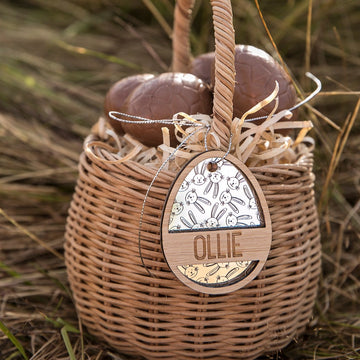 A wicker basket with straw holds two chocolate eggs. Hanging from the handle is a Personalised Golden Egg Easter Tag—an egg-shaped wooden tag engraved with OLLIE and bunny art. The basket sits on grass.