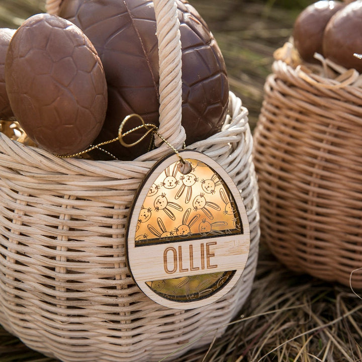 A wicker basket of chocolate Easter eggs sits on grass, with a Personalised Golden Egg Easter Tag engraved with bunny and carrot designs and the name OLLIE hanging from the handle. Another similar basket is in the background.