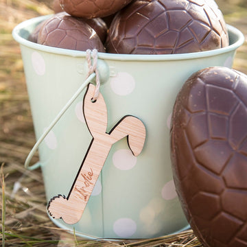 A pale green bucket with white polka dots holds large chocolate eggs, one leaning against it. An eco-friendly personalised Easter Letters tag shaped like a carrot and labeled Archie hangs from the handle.