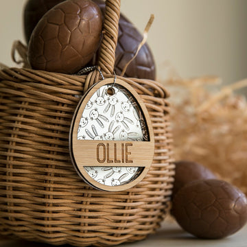 A wicker basket filled with chocolate eggs, featuring a Personalised Golden Egg Easter Tag engraved with bunny illustrations and the name OLLIE. Straw and more chocolate eggs surround the display.