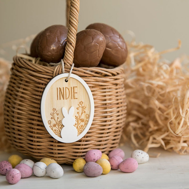 A wicker basket filled with chocolate eggs includes the Personalised Spring Fling Easter Bunny Tag, engraved with "INDIE." Mini pastel Easter eggs are displayed in front, and straw decorates the background.