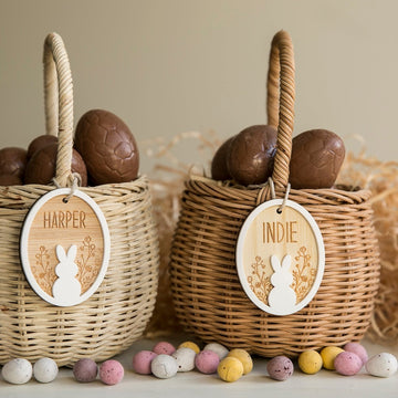 Two wicker baskets filled with chocolate eggs, each adorned with a Personalised Spring Fling Easter Bunny Tag labeled HARPER and INDIE. Colorful speckled chocolate eggs are scattered in front.