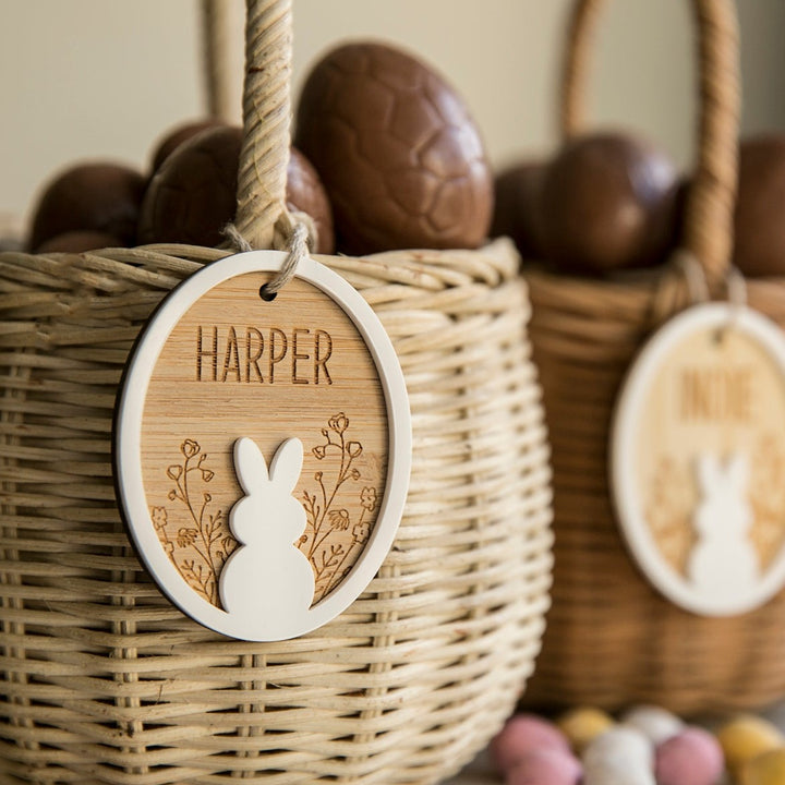 A woven basket labeled Harper with a Personalised Spring Fling Easter Bunny Tag and a bunny decoration, filled with chocolate eggs. Another similar basket is in the background, slightly out of focus, alongside pastel-colored eggs.