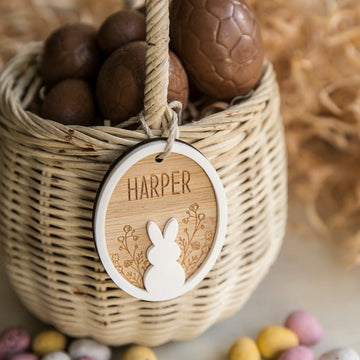 A wicker basket filled with chocolate eggs features the Personalised Spring Fling Easter Bunny Tag engraved with "Harper," floral accents, and a white bunny silhouette. Colorful mini eggs are scattered nearby.