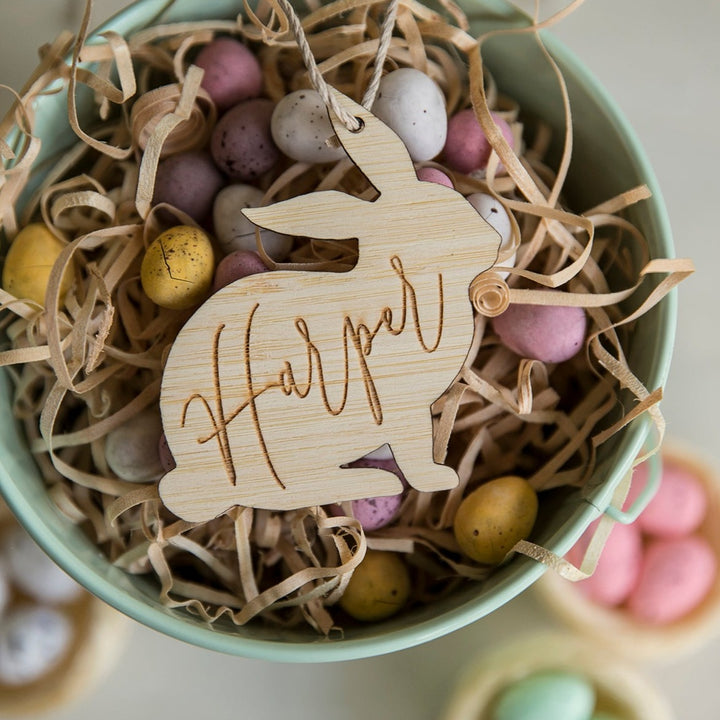 A bowl with pastel chocolate eggs and shredded paper features a Personalised Vintage Easter Bunny Tag, crafted from eco-friendly bamboo and engraved with the name Harper.