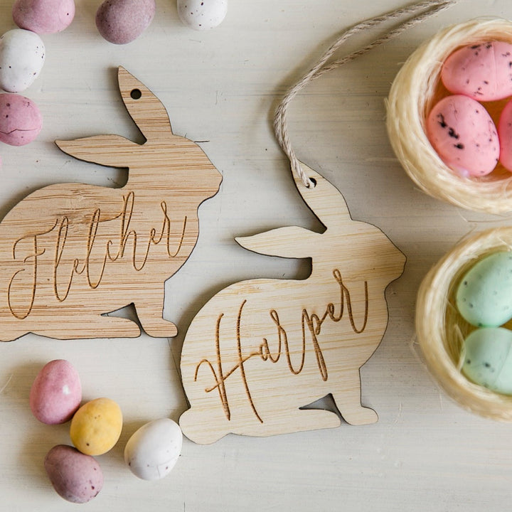 Two Personalised Vintage Easter Bunny Tags labeled Fletcher and Harper, crafted from eco-friendly bamboo, are displayed with pastel candy eggs and a basket—an ideal touch for Easter Bunny celebrations.