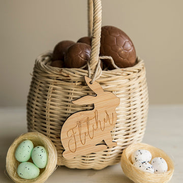 A woven bamboo basket filled with chocolate eggs is topped with a Personalised Vintage Easter Bunny Tag inscribed "Fletcher," and sits beside two nests, one with green eggs and one with white eggs.