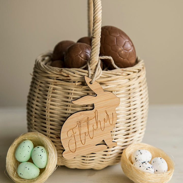 A woven bamboo basket filled with chocolate eggs is topped with a Personalised Vintage Easter Bunny Tag inscribed "Fletcher," and sits beside two nests, one with green eggs and one with white eggs.