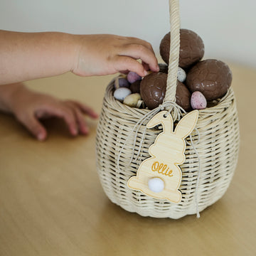 A child’s hand reaches for chocolate eggs in a basket adorned with a Personalised Easter Bunny Tag, featuring the name “Ollie,” on a wooden surface.
