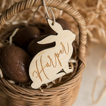 A wicker basket with chocolate eggs and a Personalised Vintage Easter Bunny Tag, crafted from eco-friendly bamboo and engraved with the name Harper, surrounded by straw-like material—ideal for your little Easter Bunny.
