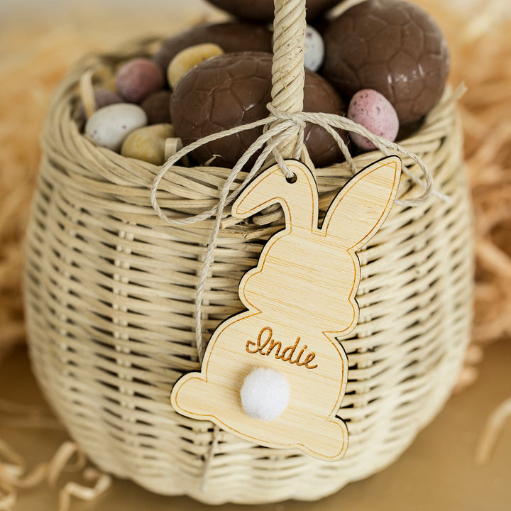 A wicker basket holds chocolate eggs and mini speckled candies. The handle is tied with a Personalised Easter Bunny Tag featuring a white pom-pom tail and the name Indie.